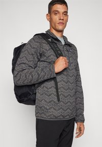 Gray patterned fleece jacket with a zippered collar, worn over a light shirt, paired with a black backpack featuring adjustable straps.