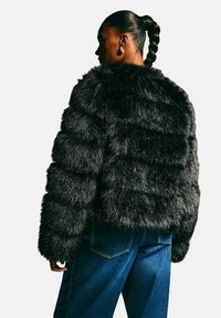 Mujer con coleta trenzada, vistiendo grandes pendientes de aro, una chaqueta de piel sintética negra y jeans azules, de pie de lado frente a un fondo liso.