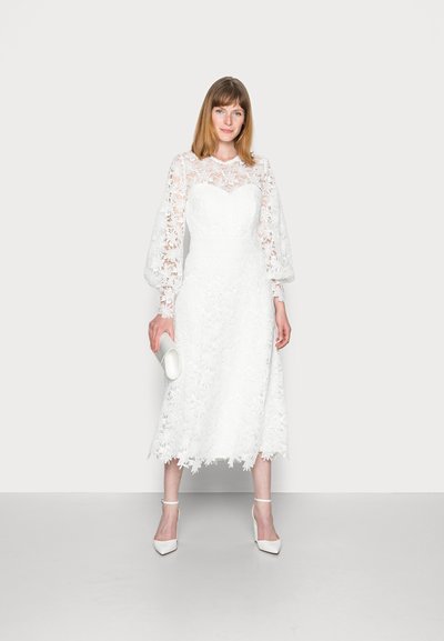 Witte kanten midi-jurk met bloemendetails, doorschijnende lange mouwen en een getailleerd lijfje, gecombineerd met een witte clutch en sandaaltjes.