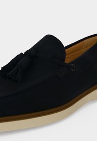 Mocassin en daim noir avec détail pompon, doublure intérieure beige et semelle blanche en caoutchouc.