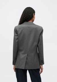 Blazer gris con rayas en espiga y una abertura en la parte posterior, corte entallado, botones negros contrastantes en las mangas y hombreras para mayor estructura.