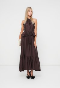 Maxikleid aus dunkelbraunem Stoff, mit Neckholder-Design, Peplum-Taille, geschichtetem Rüschensaum, getragen mit einer weißen Clutch und schwarzen Schleifenabsätzen.
