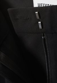 Close-up of black pants waistband showing two silver hook-and-bar fasteners and partially visible zipper with "WEEKDAY" label.