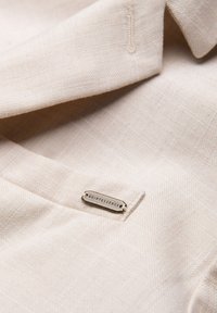 Tissu beige texturé avec un col et une étiquette métallique portant la mention « Quintessence » attachée près de la couture.