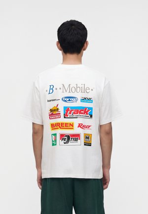 Homme portant un t-shirt blanc avec divers logos colorés et textes sur le thème de la course et des moteurs dans le dos, debout devant un fond uni.