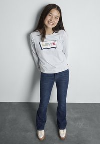 Svetlomodrá košeľa s dlhým rukávom a viacfarebným logom "Levi's", skombinovaná s tmavými rozšírenými džínsy a bielymi teniskami s hnedými podrážkami.
