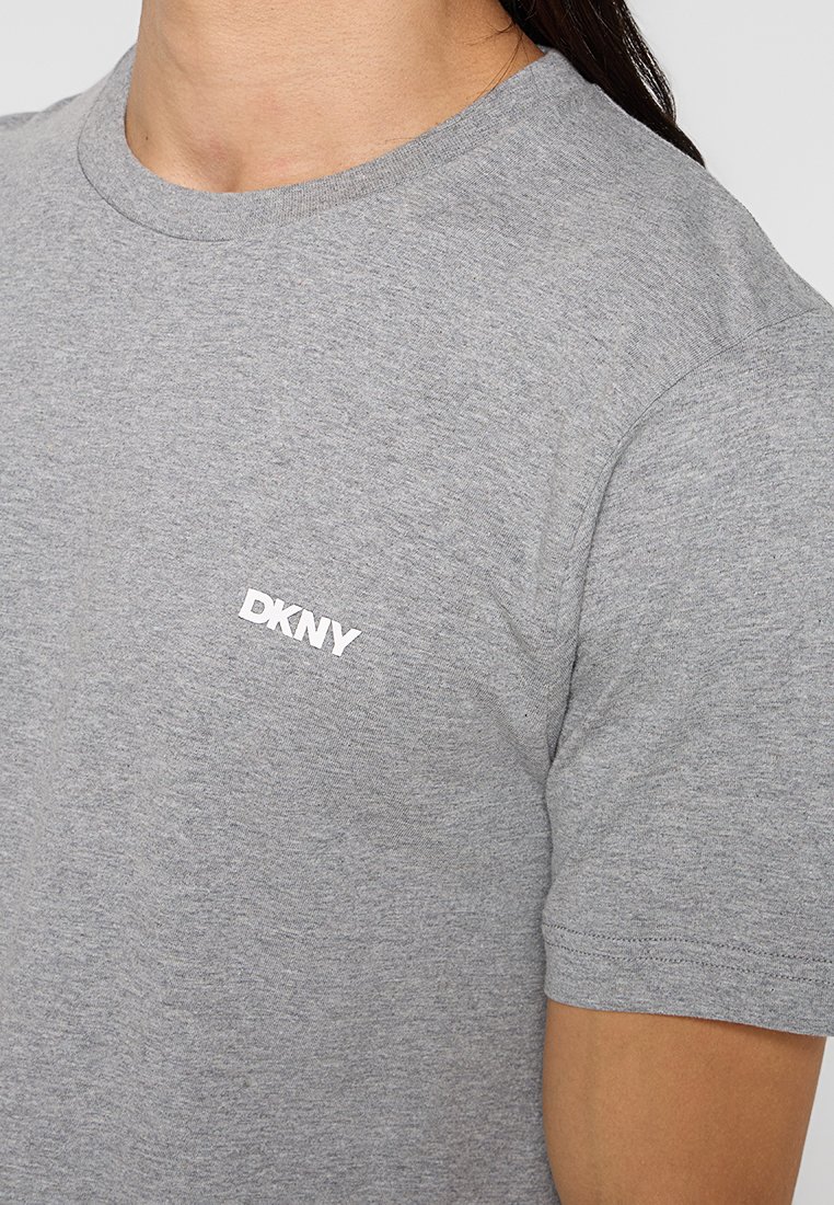 Camiseta de algodón gris con cuello redondo y mangas cortas, que presenta un logo blanco de DKNY en la parte superior izquierda. Texto suave con un ajuste casual.