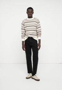 Un pull en tricot rayé crème clair et marron associé à un jeans droit noir et des baskets beiges avec des semelles marron, positionné sur un fond blanc.