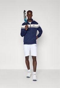 Marine zip-up jas met witte en blauwe strepen, witte shorts, houdt een blauw en zwart tennisracket vast, draagt witte sneakers.
