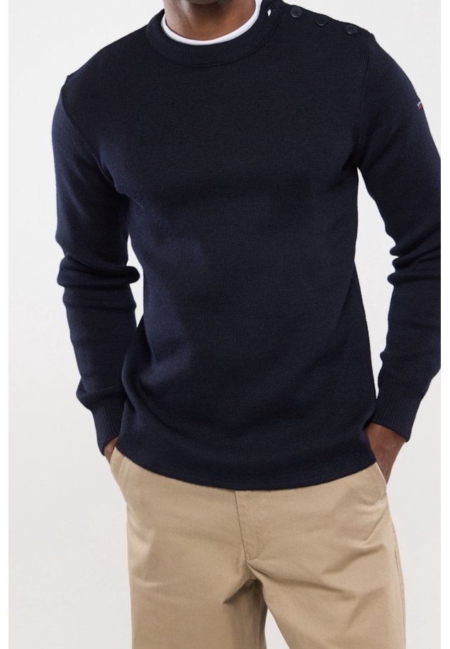 Homme portant un pull en maille bleu marine avec des boutons aux épaules et un pantalon beige, debout les mains dans les poches devant un fond uni.