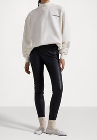 Henkilö, jolla on yllään ylisuuri valkoinen huppari, mustat nahkalegginsit, kerma-valkoiset neulotut sukat ja valkoiset balettikengät, seisomassa tasaisella taustalla.