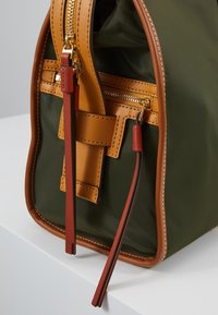Gros plan d’un sac en tissu vert avec garniture en cuir beige, fermeture éclair dorée et languettes en cuir marron sur une surface blanche.