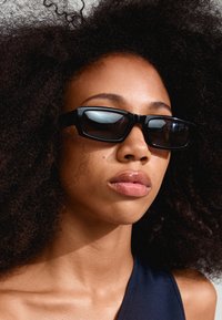 Black square sunglasses with a thick frame, dark lenses, and glossy finish; model has curly hair and natural makeup.