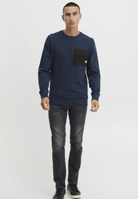 Marineblå hettegenser med en svart lomme på brystet, rund halslinning og ribbede mansjetter, kombinert med mørke jeans og svarte joggesko.