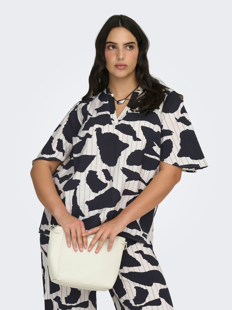 Femme aux cheveux ondulés et foncés portant un ensemble assorti top et pantalon à imprimé abstrait noir et blanc, tenant un petit sac à main blanc devant elle.