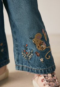 Denimjeans mit einem bestickten gelben Maus, Blumen und Blättern am Saum, kombiniert mit hellrosa Sneakers mit blumengemusterten Schnürsenkeln.