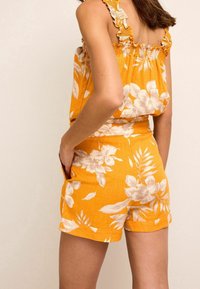 Oranssi kukkaroitu haalari, jossa on teksturoitu kangas, röyhelöolkaimet ja taskut, löysä istuvuus ja yksityiskohtaiset valkoiset kukkakuviot.