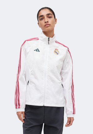 Mujer con el cabello oscuro recogido, lleva una chaqueta deportiva blanca con rayas rosas y logos de Real Madrid y Adidas, de pie frente a un fondo liso.