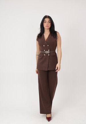 Kvinde med langt sort hår iført ærmeløs brun dobbeltradet blazer, matchende vidde bukser, et dekorativt bælte og røde spidse hæle.