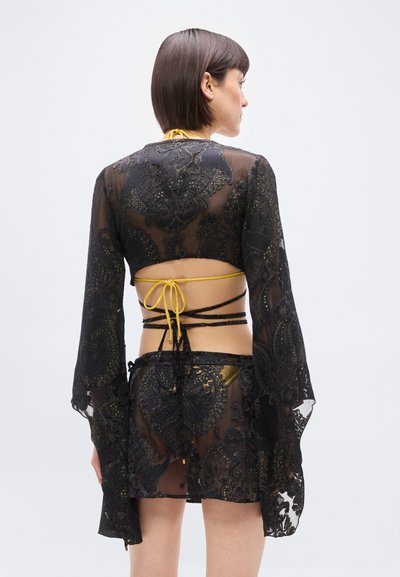 Γυναίκα με την πλάτη, φορώντας διαφανές μαύρο δαντελωτό κάλυμμα με χρυσά σχέδια πάνω από κίτρινο μπικίνι, με κορδόνια γύρω από την πλάτη και τη μέση.