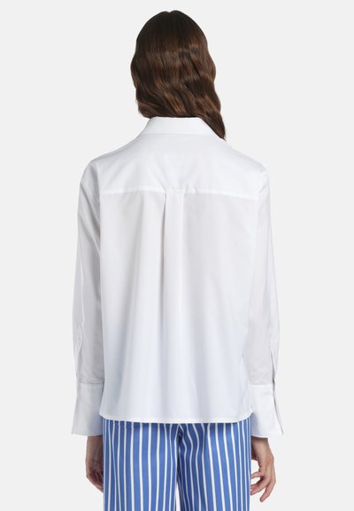 Femme aux cheveux bruns ondulés portant une chemise blanche ample à manches longues et un pantalon à rayures verticales bleu et blanc, vue de dos.