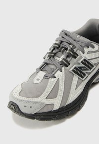Chaussure de sport grise avec tige en mesh, superpositions en synthétique et semelle extérieure en caoutchouc noir. Comprend des lacets et des accents de logo bien visibles.
