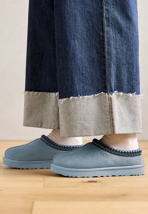 Blue suede slip-on shoes with black stitched trim worn with white socks and wide, cuffed denim jeans on a wooden floor.