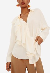 Blusa bianca con ruffles sovrapposti, chiusura a bottoni e dettaglio della cravatta regolabile. Il tessuto appare leggero e morbido. Abbinata a pantaloni marroni.