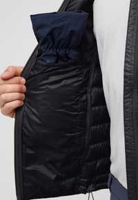 L'intérieur de la veste noire présente une doublure lisse avec une poche cachée contenant une capuche navy pliée. La fermeture éclair et la texture matelassée sont visibles.