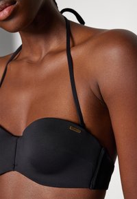 Haut de bikini noir avec un design lisse et rembourré, doté de fines bretelles et d'un petit logo doré sur le côté. Tissu doux et extensible.