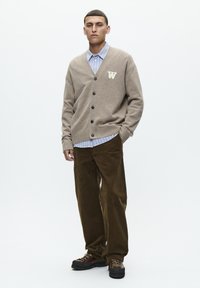 Beige cardigan med V-hals, fem knapper og et mønstret lap, båret over en lyseblå stribet skjorte, parret med mørkebrun fløjlsbukser.