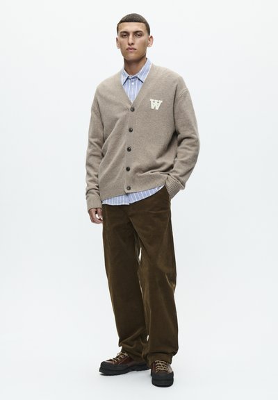 Beige cardigan with V-neck, five buttons, and a patterned patch, worn over a light blue striped shirt, paired with dark brown corduroy pants.