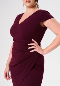 Burgunderfarbenes Kleid mit kurzen Ärmeln und einem V-Ausschnitt. Verfügt über drapierte Details an der Seite und einen glatten, dehnbaren Stoff.