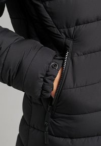 Sort puffermantel med quiltet tekstur, der har en lynlåslomme i siden, elastiske håndled og en glat overflade.