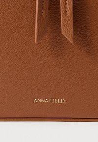 Brun lædertaske med tekstur, med klap og to lange stropper, med en gylden "ANNA FIELD"-logo på fronten.