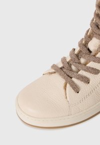 Sneaker in pelle testurizzata di un beige chiaro con spesse stringhe marroni annodate e cuciture visibili su uno sfondo bianco semplice.