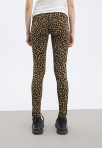 Person som bär tights med leopardmönster och svarta träningsskor, står mot en enfärgad vit och grå bakgrund.