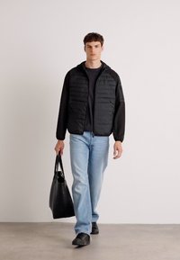 Schwarze gesteppte Jacke mit langen Ärmeln, hellblaue Jeans, schwarze Schuhe und eine schwarze Tote-Bag, vor einem neutralen Hintergrund.
