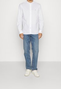 Camicia bianca a maniche lunghe con colletto alla coreana e chiusura con bottoni, abbinata a jeans in denim azzurro chiaro e sneakers bianche. Fit casual.
