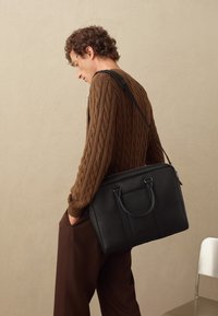 Bolso mensajero de cuero negro con asas superiores y correa para el hombro. Textura suave con costuras verticales y hardware mínimo. Suéter de punto de cable marrón.