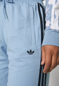 Ljusblå Adidas sweatpants med svarta ränder och logotyp, ihop med en hand som vilar vid benet som bär en blå och vit långärmad tröja.