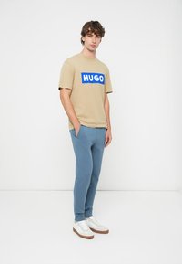 Bež pamučna majica kratkih rukava s plavim logotipom "HUGO", uparena s svijetloplavim trenirkama i bijelim tenisicama, stojeći na bijeloj pozadini.