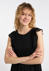 Czarna sukienka z teksturowanej tkaniny, z falbaniastymi krótkimi rękawami i okrągłym dekoltem, prezentowana na osobie z założonymi rękami.