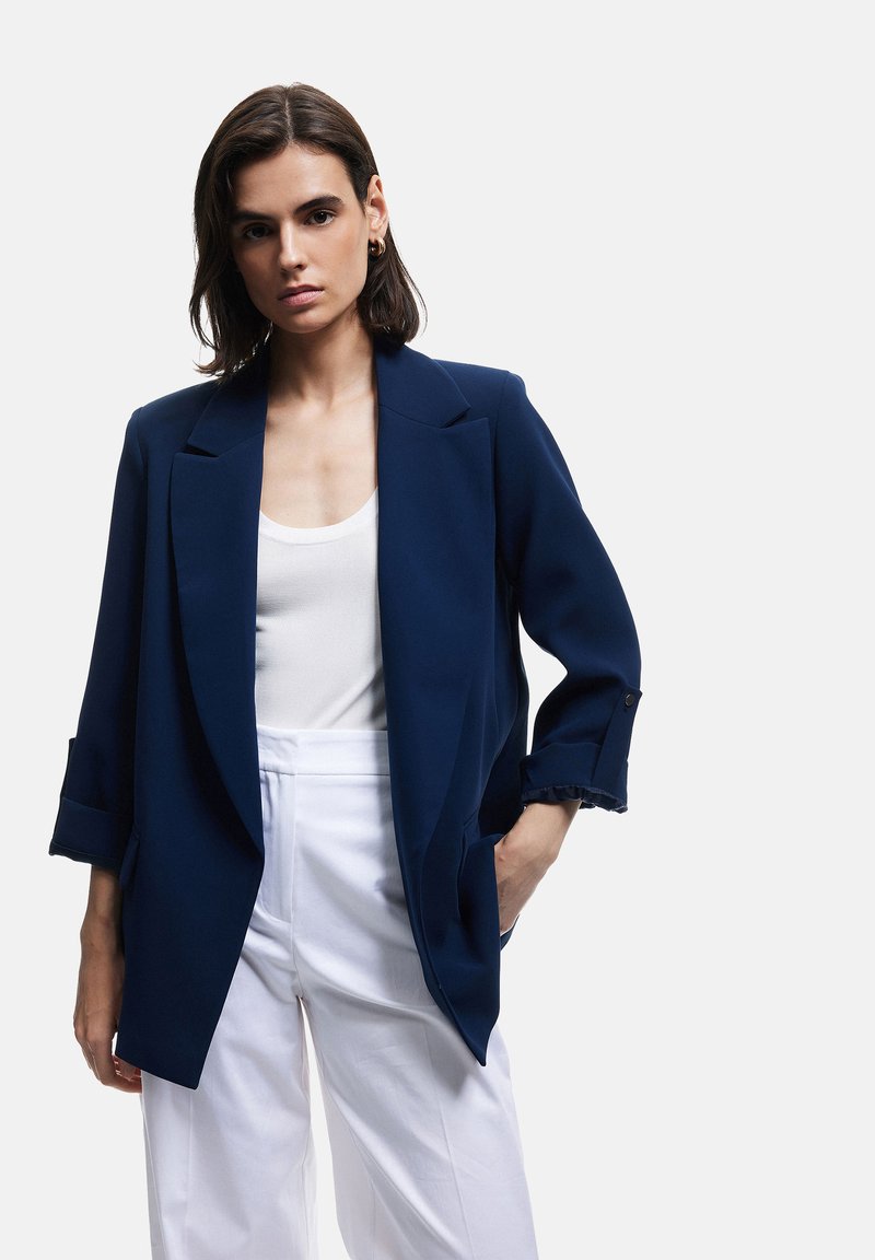 Blazer azul marino con solapas estructuradas, mangas remangadas y dos bolsillos frontales, combinado con una blusa blanca y pantalones blancos de cintura alta.