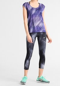 Vrouw draagt een paars, patroonrijk kort shirt, zwart-grijs patroonrijke cropped legging met Nike-logo's en teal Nike hardloopschoenen.