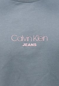 Γκρι-μπλε βαμβακερό T-shirt με την επιγραφή «Calvin Klein» σε ροζ γραφή και «JEANS» σε έντονα, αντίθετα γράμματα στο μπροστινό μέρος. Μαλακή υφή.