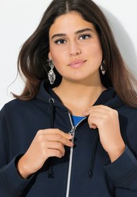 Sweat à capuche zippé bleu marine avec cordon tressé, fermeture éclair argentée, superposé à une chemise bleue. Le modèle porte des boucles d'oreilles argentées imposantes.