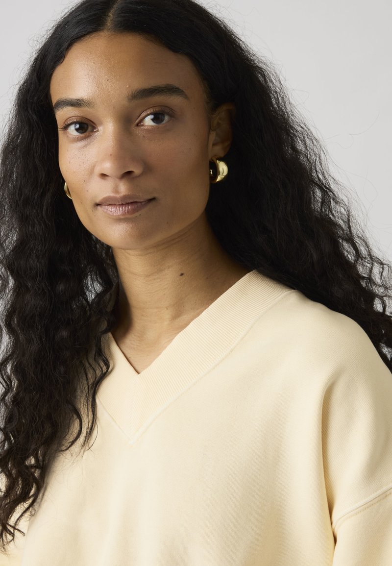 Femme aux longs cheveux bouclés et foncés, portant des boucles d'oreilles dorées en forme de créoles et un pull crème à col en V, face à l'avant avec une expression neutre.