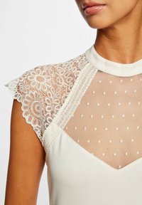 Hvid top med blonde-kapelærmer og et gennemsigtigt, prikket meshpanel ved halsudskæringen. Glat stof med blomsterblonde-detaljer.