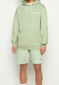 Felpa verde chiaro con tasca a marsupio e lacci bianchi. Abbinata a pantaloni corti verde chiaro coordinati, con ricamo del logo.